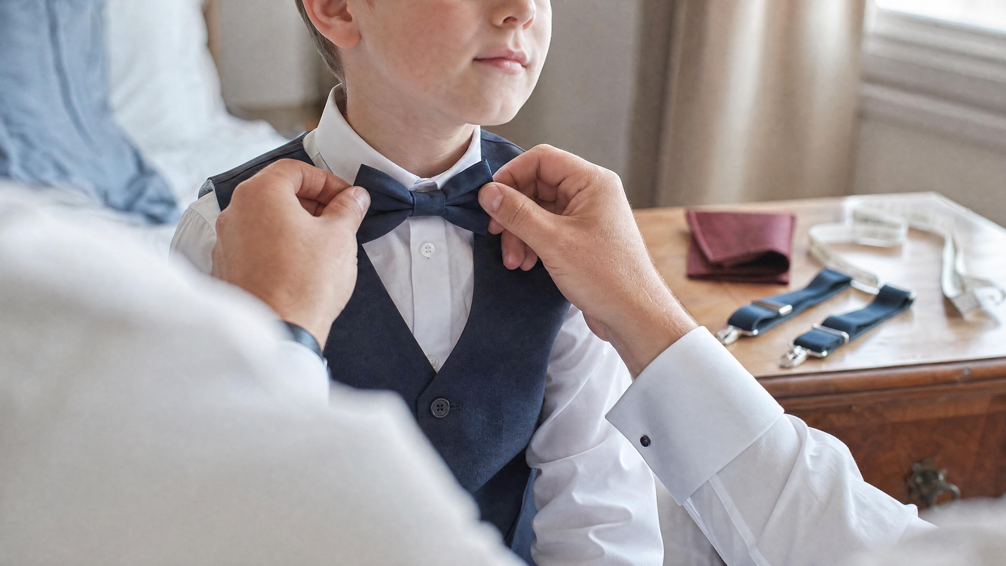 Un adulte ajuste un nœud papillon sur un enfant en chemise blanche, avec un mètre ruban visible sur une commode, dans une chambre lumineuse avant une cérémonie Kuva